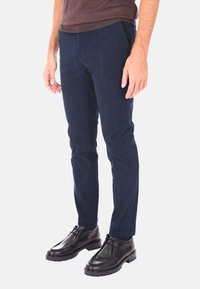 Uomo in pantaloni di velluto a coste blu navy e scarpe di pelle nere in piedi davanti a uno sfondo bianco semplice.