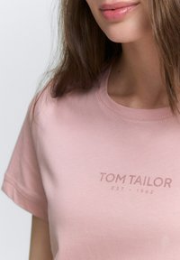 Světle růžové bavlněné tričko s kulatým výstřihem a krátkými rukávy. Text "TOM TAILOR EST - 1962" potisknutý tmavě růžovou barvou. Hladký povrch.
