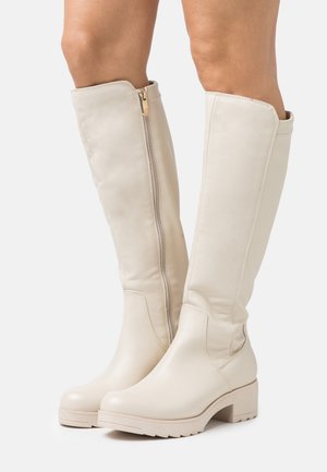 Beige knæhøje læderstøvler med sidelås og lav blokhæl, båret på bare ben mod en hvid baggrund.