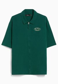 Camisa verde de manga corta hecha de algodón, con cierre frontal y cuello redondeado, con un detalle de insignia bordada.