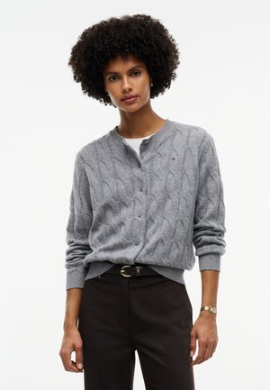 Kvinde med krøllet hår iført en grå kabelstrikket cardigan med knapper, sorte bukser med bælte og et armbåndsur på en hvid baggrund.