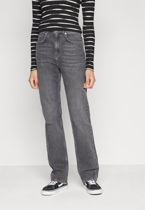 Person trägt hochgeschnittene, ausgewaschene schwarze Jeans, ein schwarz-weiß gestreiftes Langarmshirt und schwarze Sneakers mit weißen Schnürsenkeln, steht.