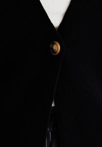 Black knitted cardigan with a v-neck design, featuring a round brown button, paired with a white ribbed top underneath.