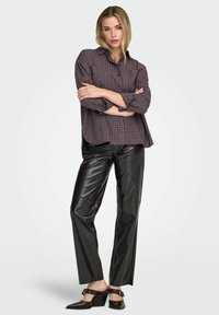 Camisa de cuadros en tonos oscuros, pantalones de cuero sintético negro brillante y zapatos oscuros de tacón alto, exhibidos con los brazos cruzados.