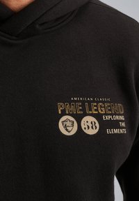 Zwarte hoodie gemaakt van zachte stof met een goud en crème graphic op de borst, inclusief een schildicoon en tekst elementen.