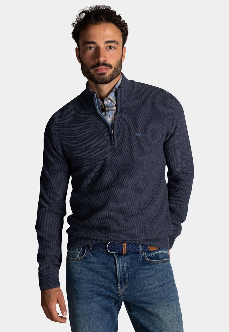 Donkerblauwe pullover met een gestructureerde tricot, een halve ritssluiting bij de kraag en contrasterende blauwe accenten. Draagt dit over een geruit overhemd met spijkerbroek.