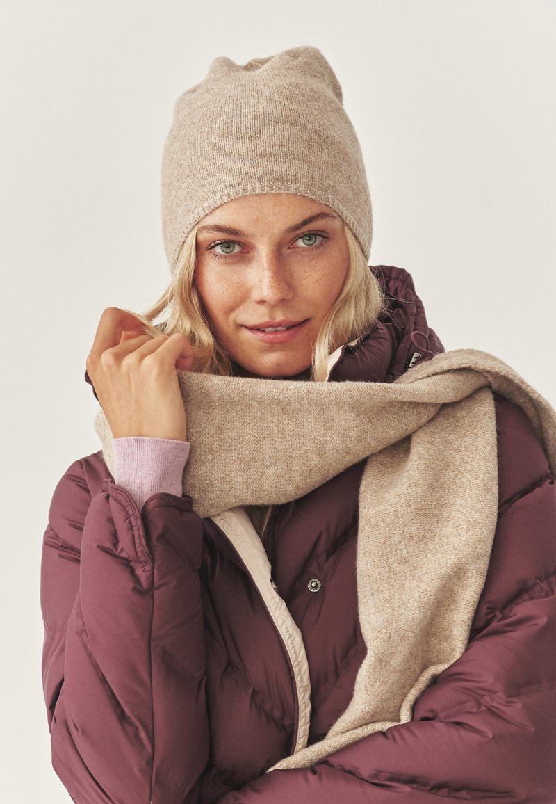 Bež pletena kapa in ujemajoč šal v paru z puhovko v globoki bordo rdeči barvi. Jakna ima diagonalne šive in lepo podlogo.