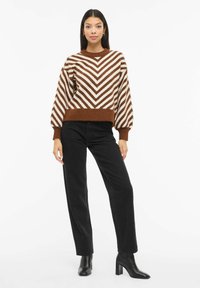 Hnědo-bílý pruhovaný svetr s dlouhými rukávy a žebrovaným lemem, kombinovaný s černými kalhotami s vysokým pasem a černými kotníkovými botami na podpatku.