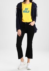 Marinblå huva jacka med dragkedja, gul bomullströja med texten "BE BOLD", svarta ribbade cropped byxor och vita slip-on skor.