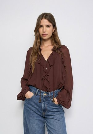 Mujer con cabello largo y castaño vistiendo una blusa marrón con volantes y vaqueros azules, de pie con las manos en los bolsillos contra un fondo liso.
