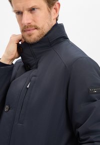 Granatowa wodoodporna kurtka z wysokim kołnierzem, dwoma guzikami, kieszenią na zamek oraz naszywką z logo na lewym rękawie. Gładka faktura.