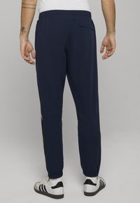 Marineblauwe lichtgewicht joggingbroek met elastische tailleband, taps toelopende pijpen, zijzakken en contrasterende zwarte zijstrepen. Draagt witte en zwarte sneakers.