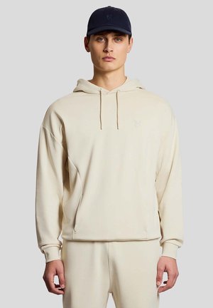 Junger Mann, der einen cremefarbenen Hoodie und passende Hosen trägt, mit einer dunkelblauen Baseballkappe, vor einem einfarbigen grauen Hintergrund stehend.