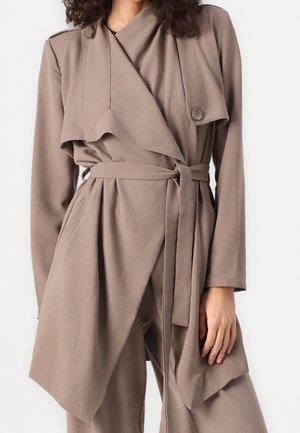 Mujer con abrigo cruzado color topo con cinturón y solapas drapeadas, combinado con pantalones de pierna ancha a juego, sobre un fondo blanco.