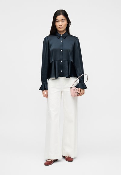 Γυναίκα που φοράει μπλε μπλούζα με κουμπιά και μανσέτες με βολάν, άσπρο παντελόνι φαρδύ, καφέ παπούτσια, κρατώντας μια μικρή ροζ τσάντα χειρός.