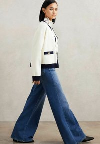 Bela pletena cardigan z mornarskimi poudarki in zlatimi gumbi, v kombinaciji s širokimi modrimi kavbojkami. Ovratnik iz čipke dodaja detajl.