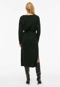 Długa, ciemnozielona dzianinowa sukienka o luźnym kroju, z wiązaniem w talii i bocznymi rozcięciami. Do niej dopasowane czarne kozaki za kolano.