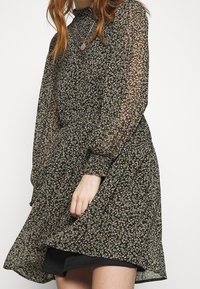 Robe noire à motifs floraux avec des manches longues transparentes, un col froncé et une taille ajustée. Le tissu a une texture délicate et légère avec de petites fleurs colorées.