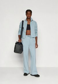 Tenue de sport bleu clair comprenant une veste zippée et un pantalon ample. Accessoires : un sac noir. Tissu lisse et coupe décontractée.