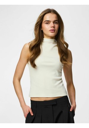 Kobieta z długimi falowanymi włosami, ubrana w białą, bezrękawnikową bluzkę z podwyższonym kołnierzem i czarne plisowane spodnie, z rękami w kieszeniach, na jednolitym tle.