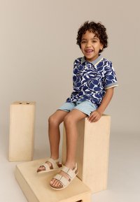 Blauwe en witte polo met patroon, lichtblauwe shorts en beige sandalen met gespen. De sandalen hebben een gestructureerde zool voor grip.