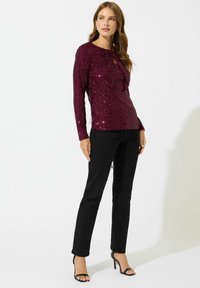 Top in paillettes bordeaux con maniche lunghe e dettaglio a occhio di bottone, abbinato a pantaloni neri aderenti e tacchi neri. Texture complessivamente liscia.