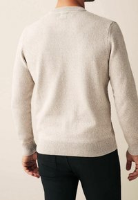 Mann, der einen hellbeigen Strickpullover und schwarze Hosen trägt, von hinten vor einem schlichten beige Hintergrund gezeigt.