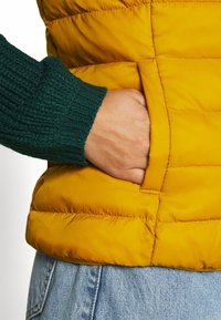 Giacca gialla imbottita con maniche lavorate a maglia verde, dotata di una tasca laterale e dettagli delle cuciture verticali sul corpo.