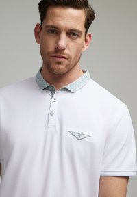 Biała koszulka polo z wzorzystym jasnoniebieskim kołnierzykiem, trzema guzikami i małą kieszonką z podobnym detalem. Teksturowana tkanina.