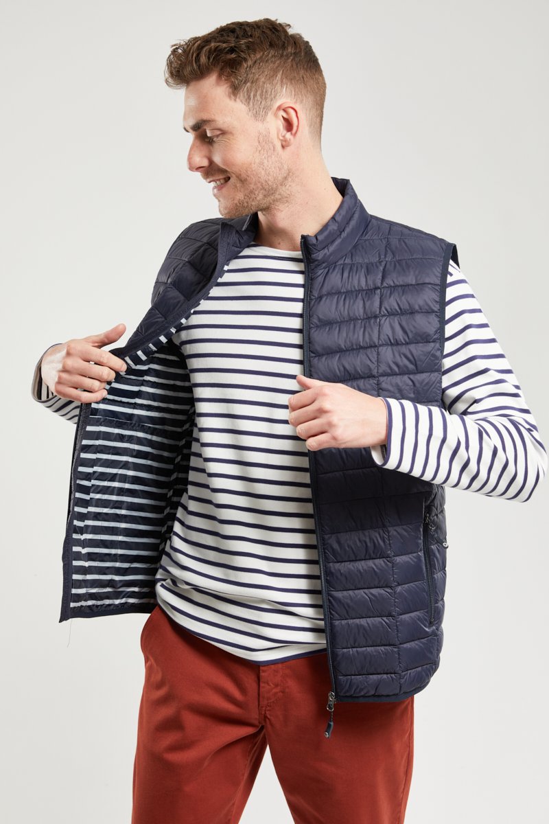 Homme portant une veste matelassée bleu marine, la dézippant pour révéler une doublure intérieure rayée, une chemise à manches longues rayée et un pantalon couleur rouille.