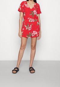 Tuta floreale rossa con maniche corte, scollo a V e vita elastica. Accentuata da fiori rosa e foglie verdi. Abbinata a sandali neri slip-on.