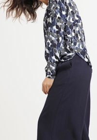 Blusa con stampa floreale in navy, verde e bianco, con maniche lunghe e vestibilità ampia, abbinata a pantaloni navy a vita alta. Tessuto morbido.