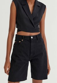 Blazer nero corto con scollatura a V profonda e design senza maniche. Abbinato a pantaloni shorts in denim neri a vita alta con bordi sfrangiati e dettagli in rame.