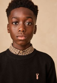 Črn pleten pulover z roza vezenim logotipom jelena, slojen preko rjave karo srajce z ovratnikom na gumbe.