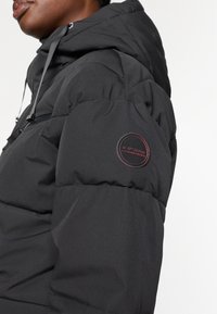 Mustard värvi paks jope, millel on tekstuurne pind, kapuuts ja ümmargune logo detail varrukal. Kujunduses on ka tepitud sektsioonid.