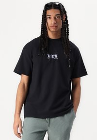 Tricou din bumbac negru cu un design grafic violet pe piept, asociat cu pantaloni cu dungi verzi. Mâneci scurte și croială lejeră.