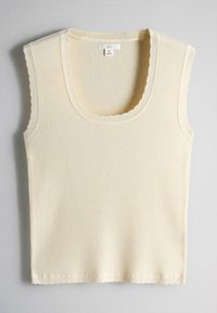 Krémovo sfarbený rebrovaný pletený bezrukávnik so zvlnenými okrajmi a výstrihom do lodičky, veľkosť stredná, položený na sivom povrchu.