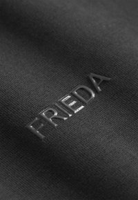 Zwarte stof met een gestructureerd oppervlak heeft het reliëfwoord "FRIEDA" in een glanzende, verheven afwerking. Geen extra patronen of accenten.