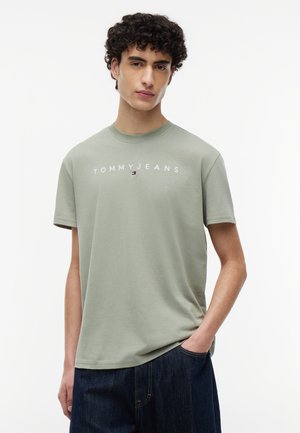 Bărbat tânăr cu păr creț și închis la culoare, purtând un tricou Tommy Jeans verde deschis și blugi albastru închis, pozând cu o mână în buzunar.