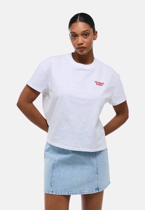 T-shirt blanche en coton à manches courtes, avec "LAS CHICAS DE IBIZA" brodé en rouge sur la poitrine. Associée à une jupe en denim bleu clair.