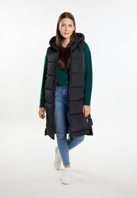 Schwarze Pufferweste mit Kapuze, mit einem gesteppten Design, Reißverschluss und seitlichen Taschen, kombiniert mit einem grünen Strickpullover und blauen Jeans.