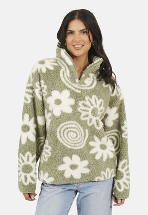 Frau trägt einen grünen Fleece-Pullover mit weißen Blumen- und Spiralmustern, kombiniert mit hellblauen Jeans und lächelt mit lockigem dunklem Haar.
