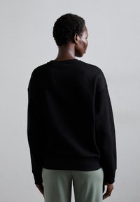Zwarte sweatshirt met een ronde hals, verlaagde schouders en geribbelde manchetten, gecombineerd met lichtgroene broek. De textuur lijkt zacht en ontspannen.