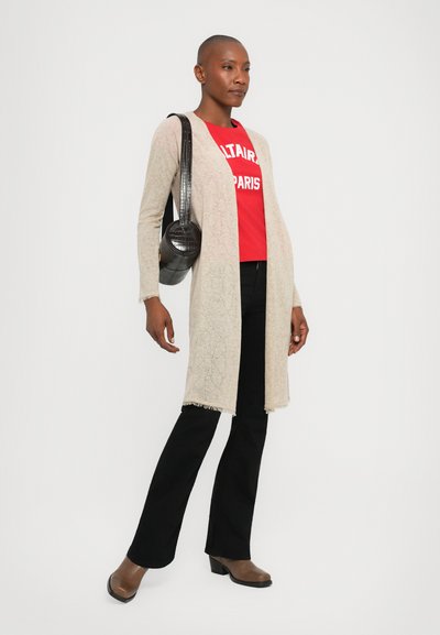 Beige long cardigan with delicate pattern, worn over a red graphic tee, paired with black flared pants and brown heeled shoes. Black circular handbag.