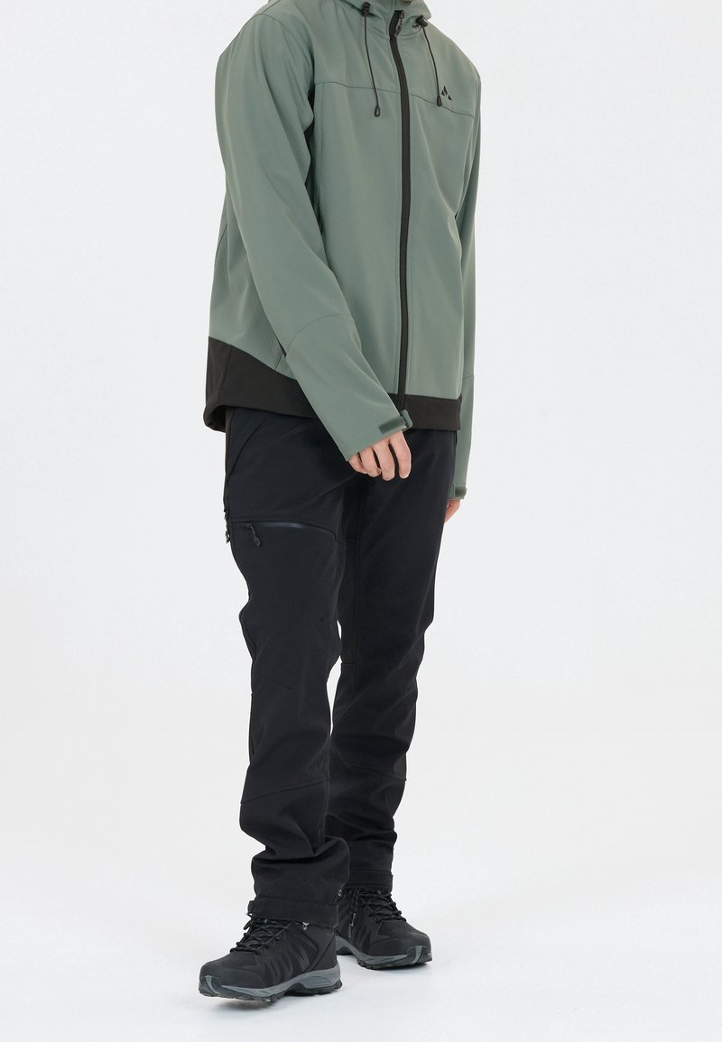 Veste softshell verte pour homme avec panneau bas noir, fermeture éclair à l'avant et capuche avec cordon, associée à un pantalon noir et des bottes noires robustes.