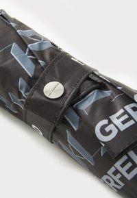 Housse de parapluie noire fabriquée en tissu durable, présentant un motif géométrique et une fermeture à bouton argenté avec détail de logo.
