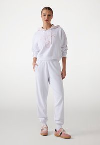 Witte katoenen hoodie met roze accenten, voorzien van een logo en een trekkoord. Gecombineerd met bijpassende witte sweatpants en roze sneakers met zwarte strepen.