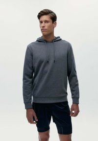 Sivá mikina s kapucňou vyrobená z mäkkej tkaniny, s prednou vreckom, kapucňou na šnúrku a rebrovanými manžetami, v kombinácii s čiernymi šortkami.