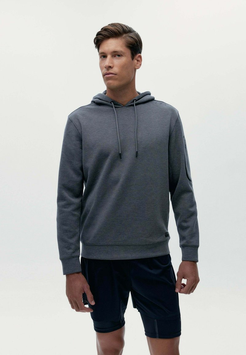 Sivá mikina s kapucňou vyrobená z mäkkej tkaniny, s prednou vreckom, kapucňou na šnúrku a rebrovanými manžetami, v kombinácii s čiernymi šortkami.