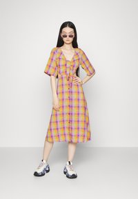 Vestido tipo camisa, até ao joelho, em xadrez multicolorido de rosa, laranja e roxo. Apresenta mangas curtas em forma de balão e um detalhe de laço na frente.